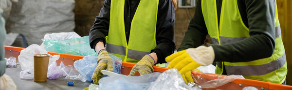 Cropped view of sorters in high visibility vests and gloves sorting garbage on conveyor while working in waste disposal station at background, garbage sorting and recycling concept, banner 