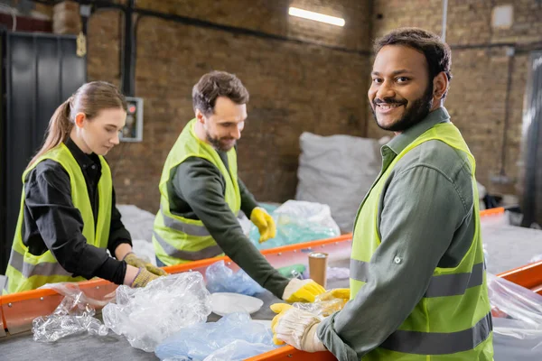 Joyful indian worker in protective vest and gloves looking at camera while standing near trash on conveyor and blurred colleagues in garbage sorting center, garbage sorting and recycling concept