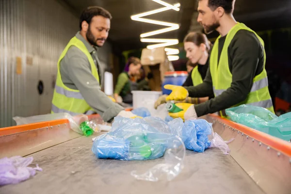 Plastic trash on conveyor near blurred multiethnic workers in high visibility vests and gloves working together in garbage sorting center, garbage sorting and recycling concept