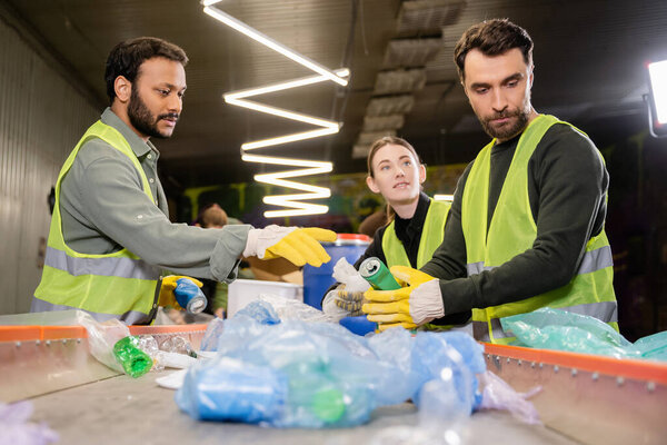 Мультиэтнические сортировщики в защитной одежде и перчатках беседуют с коллегами при работе с пластиковым мусором на конвейере на станции утилизации отходов, концепции сортировки и переработки мусора