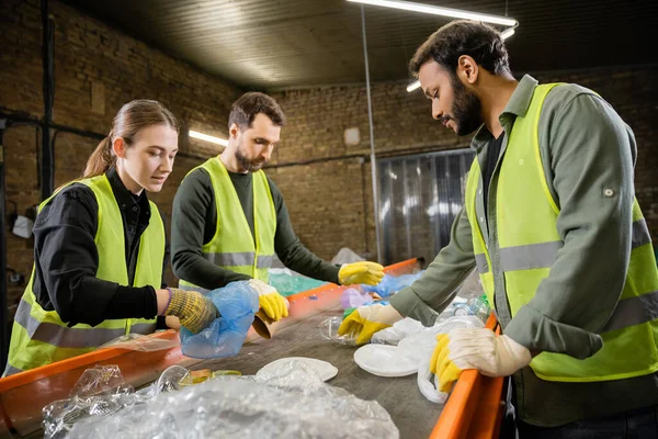 stock image Multiethnic workers in protective vests and gloves taking trash from conveyor while working together in waste disposal station at background, garbage sorting and recycling concept