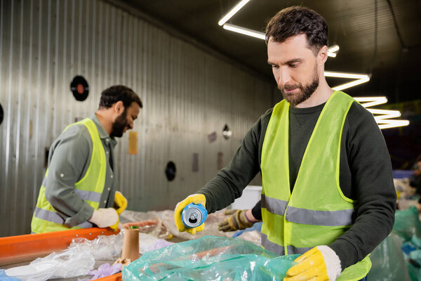 worker in protective gloves and vest putting tin can in plastic bag while separating trash near multiethnic colleagues and conveyor in waste disposal station, garbage sorting and recycling concept