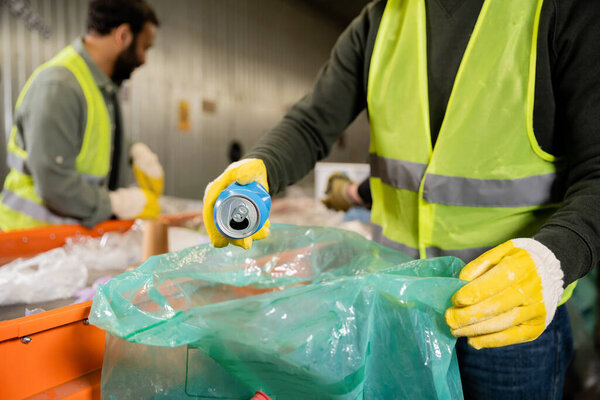 Cropped view of man in protective vest and gloves putting tin can in plastic bag while working near blurred multiethnic colleagues and conveyor in waste disposal station, recycling concept