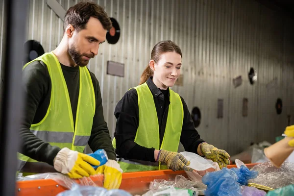 在废物处置站与同事一起工作时 穿着防护手套和安全背心在传送机上对垃圾进行分类的年轻工人微笑 回收概念 — 图库照片