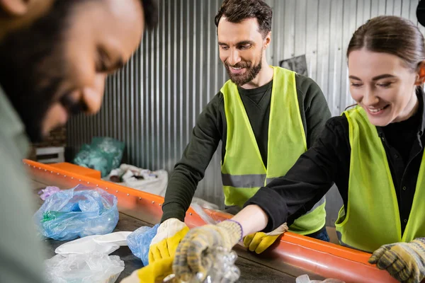 Smiling and bearded worker in protective vest and gloves taking and separating trash on conveyor near cheerful interracial colleagues working in garbage sorting center, recycling concept