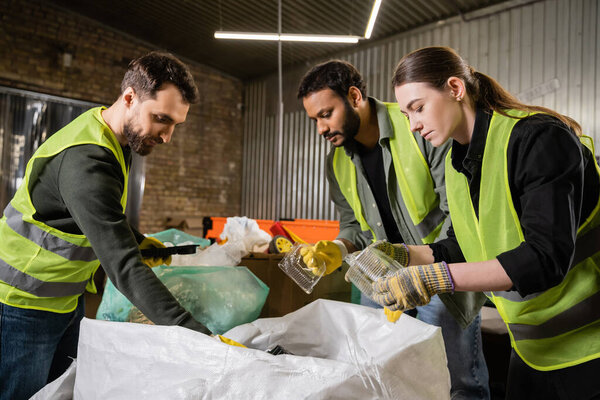Interracial sorters in protective gloves and safety vests taking plastic containers from sacks while sorting trash together in waste disposal station, garbage sorting and recycling concept