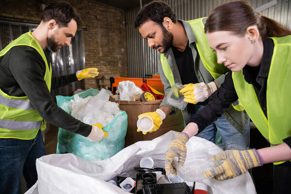 Indian sorter in high visibility vest and gloves holding plastic trash for recycling near sack while working with colleagues together in waste disposal station, garbage sorting and recycling concept