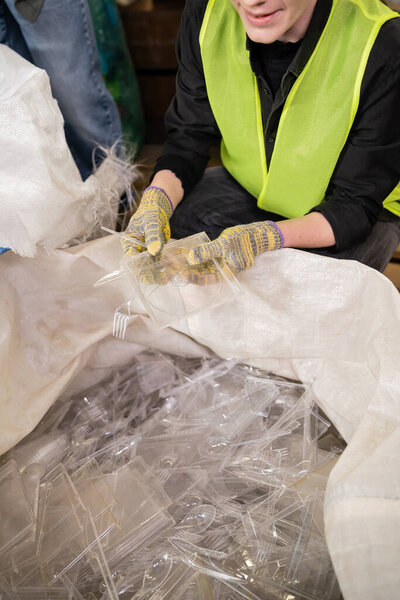 Cropped view of smiling female sorter in protective vest and gloves holding plastic garbage near sack while working in waste disposal station, garbage sorting and recycling concept