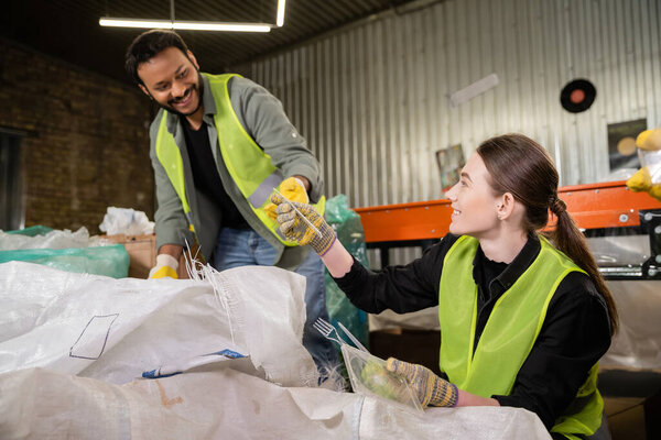 Smiling young sorter in gloves and protective vest giving plastic garbage to blurred indian colleague while working near sack in blurred waste disposal station, garbage sorting and recycling concept
