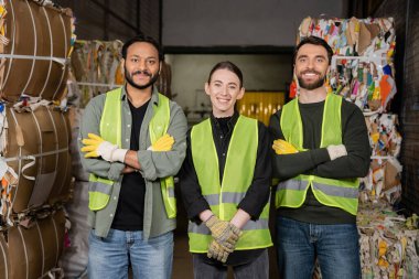 Cheerful multiethnic workers in high visibility vests and gloves crossing arms and looking at camera while standing near waste paper on disposal station, garbage sorting process  clipart