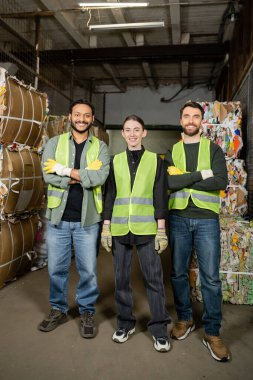 Smiling and interracial workers in high visibility vests and gloves crossing arms and looking at camera while standing near waste paper on disposal station, garbage sorting process  clipart