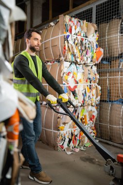 Bearded male worker in protective vest and gloves using hand pallet truck while standing near waste paper in garbage sorting center, waste sorting and recycling concept clipart