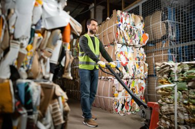 Bearded sorter in protective gloves and vest moving waste paper on hand pallet truck in blurred garbage sorting center, waste sorting and recycling concept clipart