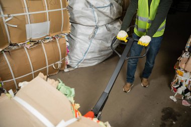 Cropped view of sorter in high visibility vest and gloves using hand pallet truck and moving waste paper in garbage sorting center, waste sorting and recycling concept clipart