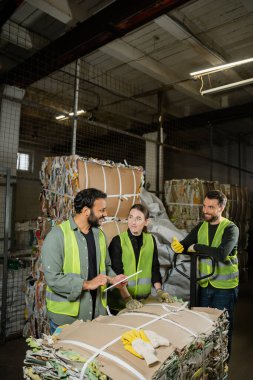 Positive indian worker in protective vest using digital tablet and talking to colleagues while standing near waste paper on hand pallet truck in waste disposal station, garbage sorting concept clipart