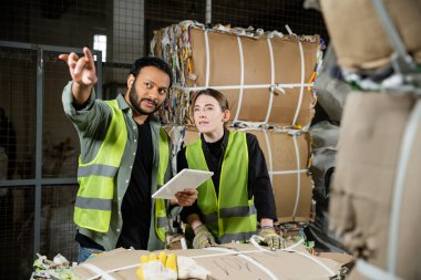 Indian worker in high visibility vest pointing with finger and using digital tablet near female colleague and waste paper on hand pallet truck waste disposal station, garbage recycling concept clipart