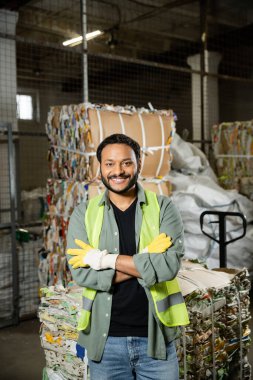 Smiling and bearded worker in protective gloves and vest crossing arms and looking at camera while standing near blurred waste paper in waste disposal station, garbage sorting and recycling concept clipart