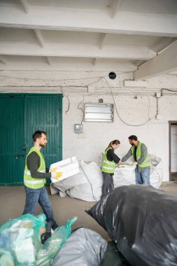 Yüksek görüş mesafesi olan yelek ve eldiven giyen erkek işçiler köpük plastik taşırken, ırklar arası meslektaşlar çöp öğütme istasyonunda çuvallarla, çöp ayıklama ve geri dönüşüm konseptiyle çalışıyorlar.