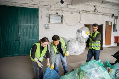 Koruyucu yelek ve eldiven giyen çok ırklı işçiler atık imha istasyonunda meslektaşlarının yanında plastik poşetlerle çalışıyorlar. Çöp tasnif ve geri dönüşüm konsepti.