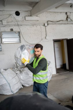 Çöp öğütme istasyonu, çöp ayıklama ve geri dönüşüm konseptinde çalışırken plastik poşet taşıyan koruyucu yelek ve eldiven giyen sakallı işçiler