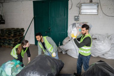 Yüksek görünürlüklü yelek ve eldiven giyen çok ırklı işçiler atık imha istasyonunda, çöp tasnif ve geri dönüşüm konseptinde meslektaşlarının yanında plastik poşetlerle çalışıyorlar.