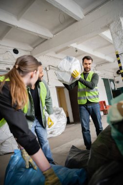 Worker in protective vest and glove holding plastic bag with trash near blurred interracial colleagues working in waste disposal station, garbage sorting and recycling concept clipart