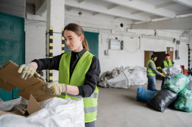 Young female worker in protective vest and gloves holding cardboard near sack and blurred colleagues working in waste disposal station, garbage sorting and recycling concept clipart