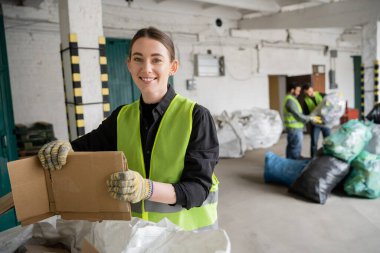 Cheerful young worker in protective vest and gloves holding cardboard and looking at camera while working near sack and blurred colleagues in garbage sorting center, recycling concept clipart