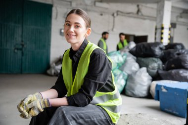 Güvenlik yeleği ve eldivenli neşeli genç işçi dinlenirken ve çöp toplama merkezinde bulanık plastik torbaların yanında otururken kameraya bakıyor, geri dönüşüm konsepti