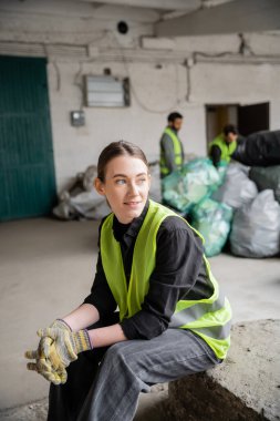 Yüksek görüş açısına sahip yelek ve eldivenlerle arka planda bulanık meslektaşları ve plastik torbaları rahatlatırken geri dönüşüm konseptini değiştiren pozitif bir genç ayıklayıcı.