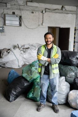 Koruyucu yelek giymiş neşeli ve sakallı Hintli işçiler, kollarını kavuşturup kameraya bakarken çöp toplama merkezinde plastik poşetlerin yanında, geri dönüşüm konseptinde