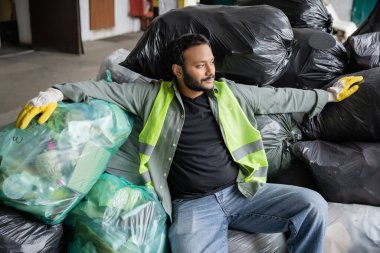 Yüksek görüş açısına sahip yelekli ve eldivenli sakallı Hintli ayırıcı çöp ayıklama merkezindeki çöp poşetlerinin üzerinde dinlenirken başka yöne bakıyor, geri dönüşüm konsepti