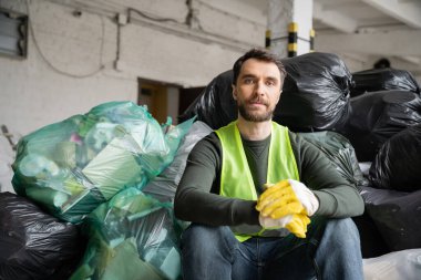 Koruyucu yelek ve eldivenli sakallı ayırıcı, çöp torbalarıyla plastik poşetlerin yanında otururken aynı zamanda bulanık çöp ayıklama merkezinde çalışırken, geri dönüşüm konseptinde çalışırken kameraya bakıyor.
