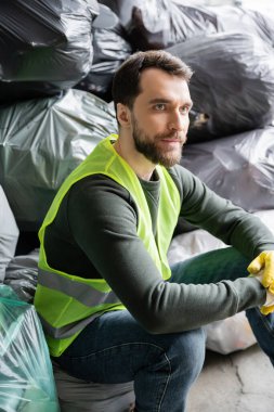 Sakallı, floresan yelekli ve eldivenli erkek işçi, çöp ayıklama merkezinde çöp dolu bulanık plastik torbaların yanında otururken başka tarafa bakıyor, geri dönüşüm konsepti