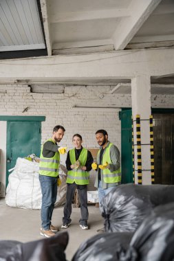 Koruyucu yelek giymiş sakallı ve eldivenli işçiler, çöp toplama merkezindeki ırklar arası gülümseyen meslektaşlar ve geri dönüşüm konseptindeki bulanık plastik torbaları işaret ediyor.