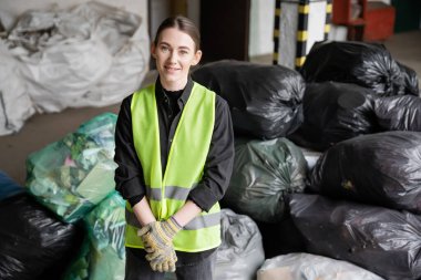 Yansıtıcı yelek ve koruyucu eldivenlerle kameraya bakarken çöp ayıklama merkezinde çöp dolu bulanık plastik torbaların yanında duran pozitif genç ayırıcı, geri dönüşüm konsepti