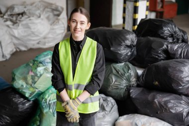 Positive young sorter in reflective vest and protective gloves looking at camera while standing near blurred plastic bags with trash in garbage sorting center, recycling concept clipart