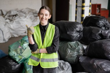 Koruyucu yelek ve eldiven giymiş gülümseyen genç işçiler jest yapıp kameraya bakarken çöp toplama merkezinde çöp dolu bulanık plastik torbaların yanında durup geri dönüşüm konseptini izliyorlar.