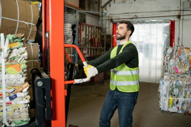 Koruyucu eldiven ve yelek sakallı ayırıcı arka planda bulanık çöp ayıklama merkezinde çalışırken atık kağıtlı el paleti kamyonu kullanıyor, geri dönüşüm konsepti