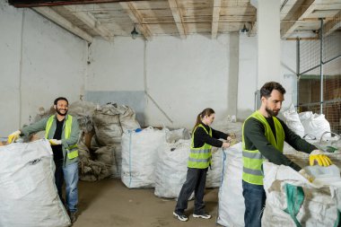 Yüksek görüş mesafesi olan yelek ve eldivenlerle gülen Hintli işçiler çöp öğütme istasyonunda, çöp ayıklama ve geri dönüşüm konseptinde, trakeoskoplu çuvallarla birlikte çalışıyorlar.