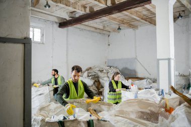 Çöp torbalarının yanında plastik çöp taşıyan koruyucu yelek ve eldiven giyen işçiler, atık öğütme istasyonu, çöp ayıklama ve geri dönüşüm kavramında çok ırklı, bulanık meslektaşlarının yanında çalışırken