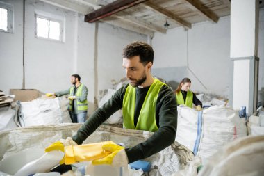 Koruyucu eldiven ve yelek giymiş sakallı bir işçi çöp torbasından plastik çöpleri alırken atık öğütme istasyonunda, çöp ayıklama ve geri dönüşüm konseptinde, bulanık meslektaşlarının yanında çalışıyor.