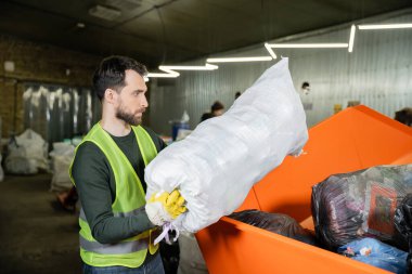 Eldiven ve koruyucu yelek giymiş sakallı ayıklayıcının bulanık atık öğütme istasyonunda çöp torbası, çöp ayıklama ve geri dönüşüm konsepti.