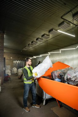 Eldiven ve floresan yelek giymiş sakallı erkek işçilerin çöp torbalarını bulanık atık öğütme istasyonunda, çöp ayıklama ve geri dönüşüm konseptinde çöp konteynerine koymalarının yan görüntüsü