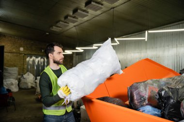 Side view of man in high visibility vest and glove putting sack with trash in container while working in waste disposal station, garbage sorting and recycling concept clipart