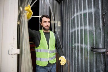 Koruyucu yelek ve eldivenli sakallı erkek işçi atık öğütme istasyonu girişinden, çöp ayıklama ve geri dönüşüm konseptine bakıyor ve yürüyor.