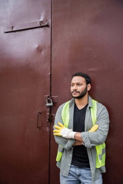 Bearded indian worker in high visibility vest and gloves crossing arms while standing near waste disposal station door, garbage sorting and recycling concept clipart