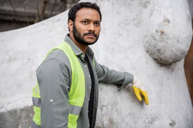 Serious indian worker in high visibility vest and glove standing near concrete sculpture in outdoor waste disposal station, garbage sorting and recycling concept clipart