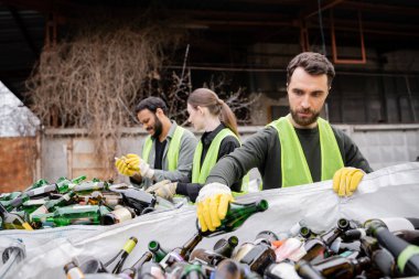 Bearded worker in protective gloves and vest putting glass bottle in sack near blurred interracial colleagues in outdoor waste disposal station, garbage sorting and recycling concept clipart