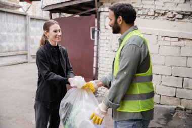 Gülümseyen genç gönüllü, atık öğütme istasyonunda, çöp tasnif ve geri dönüşüm konseptinde koruyucu yelek ve eldiven giyen Hintli işçilere çöp torbası veriyor.